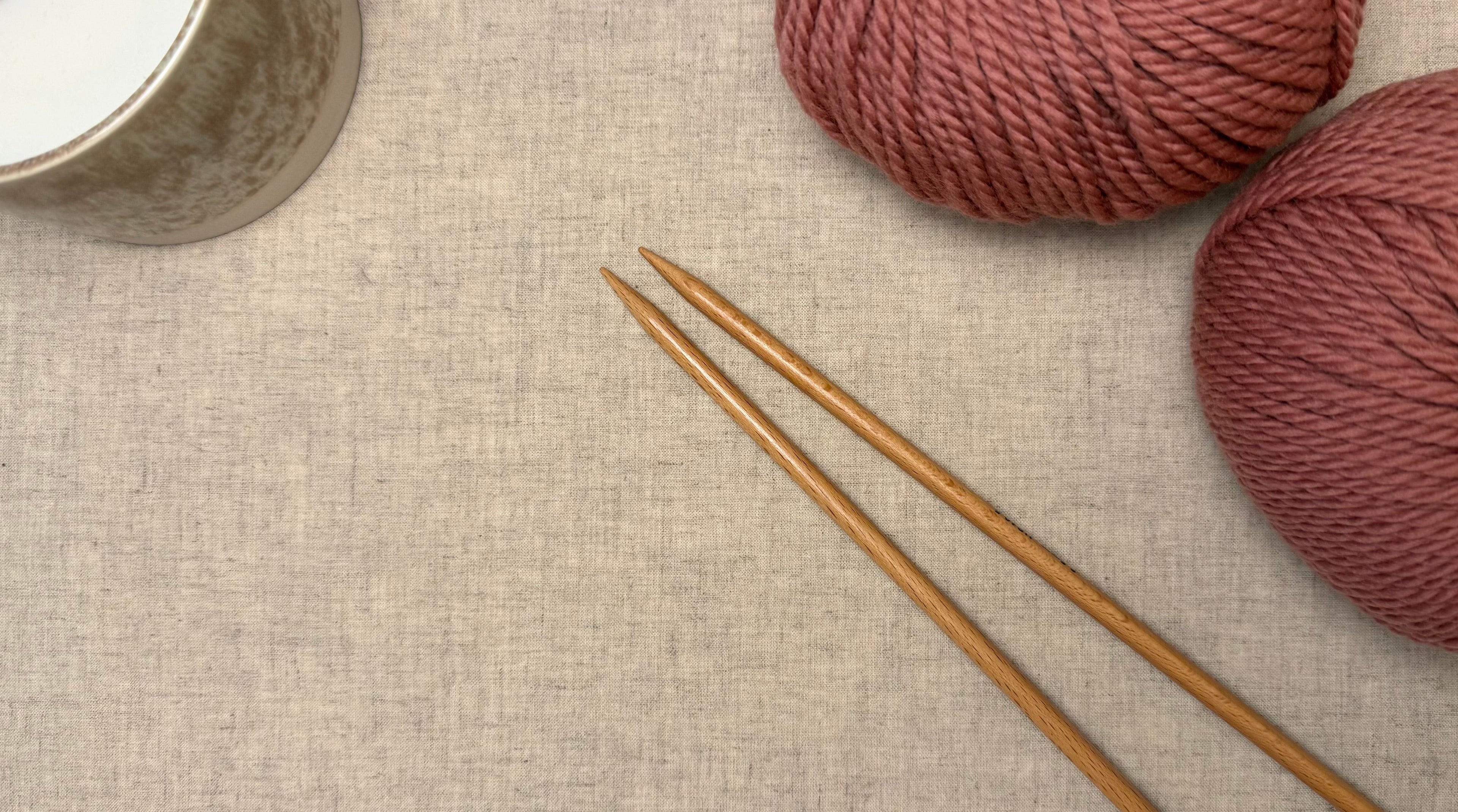 a birds eye view of some balls of yarn on a natural tablecloth with some knitting needles sitting next to them